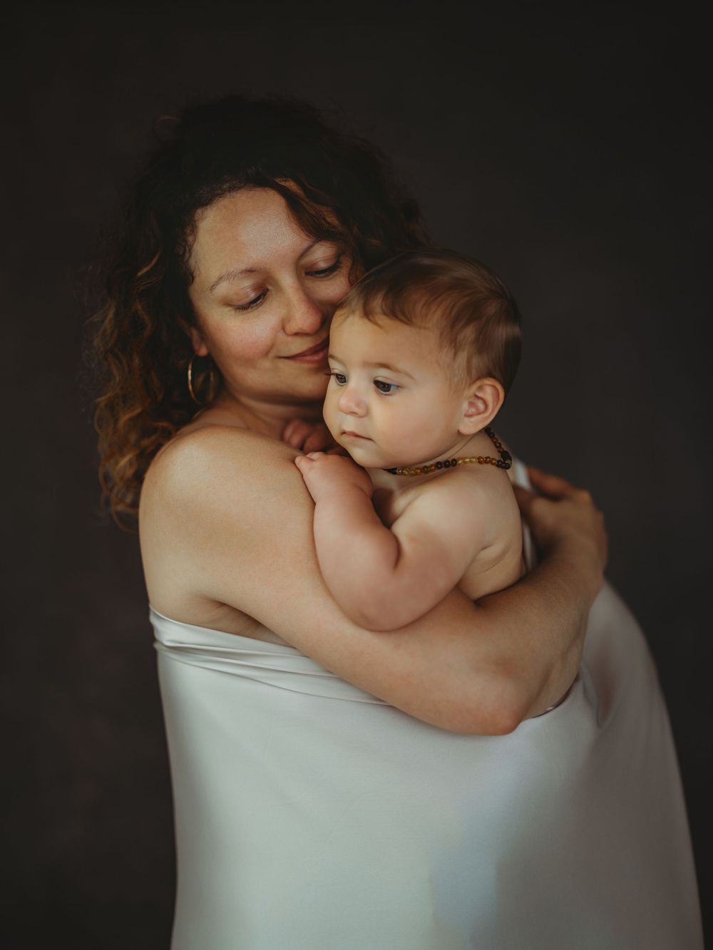 A mother with curly hair tenderly holds her baby close, both sharing a serene moment against a dark background.
