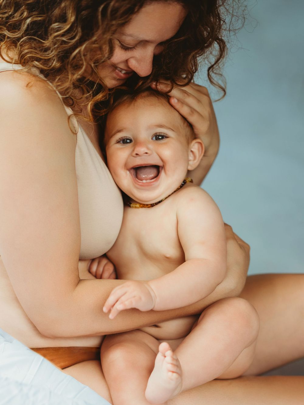 A joyful baby laughs while being held by a curly-haired mother in a warm, tender embrace against a soft blue background.