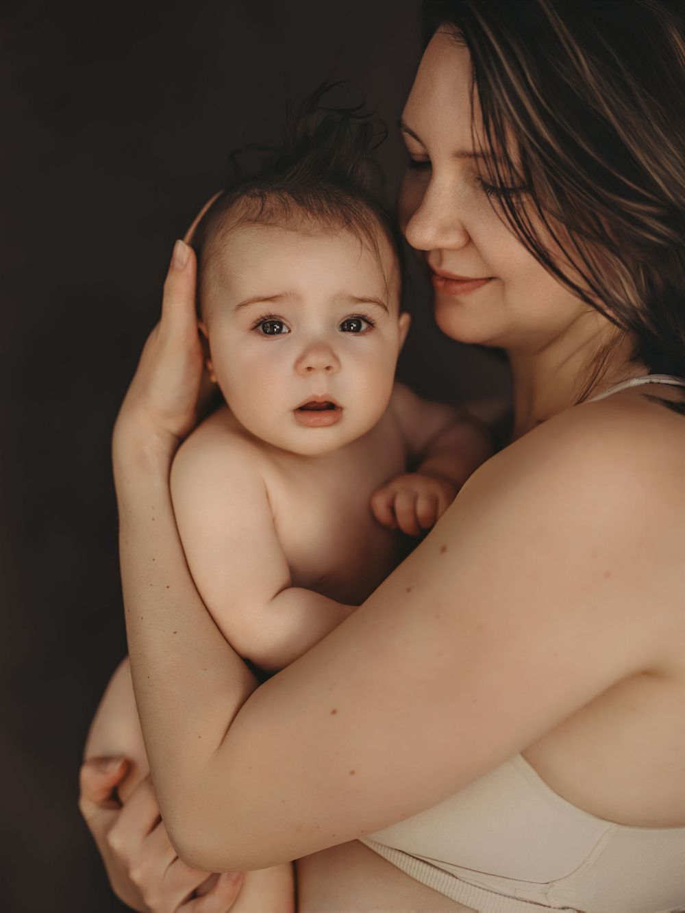 A tender moment of a mother in a beige bra cradling her wide-eyed baby against a dark studio background.