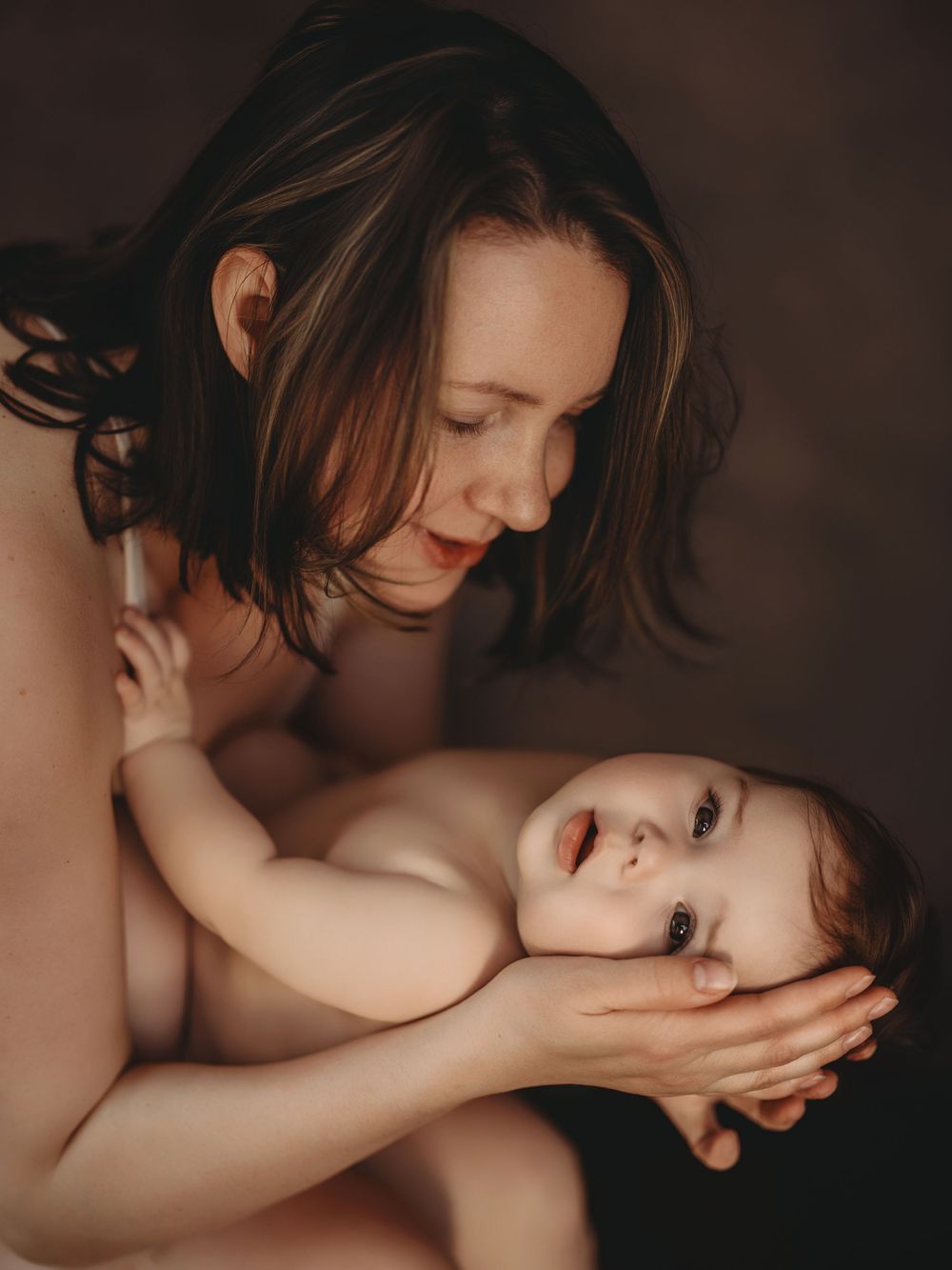 A tender moment of a mother cradling her baby, gazing lovingly at her infant against a dark background.