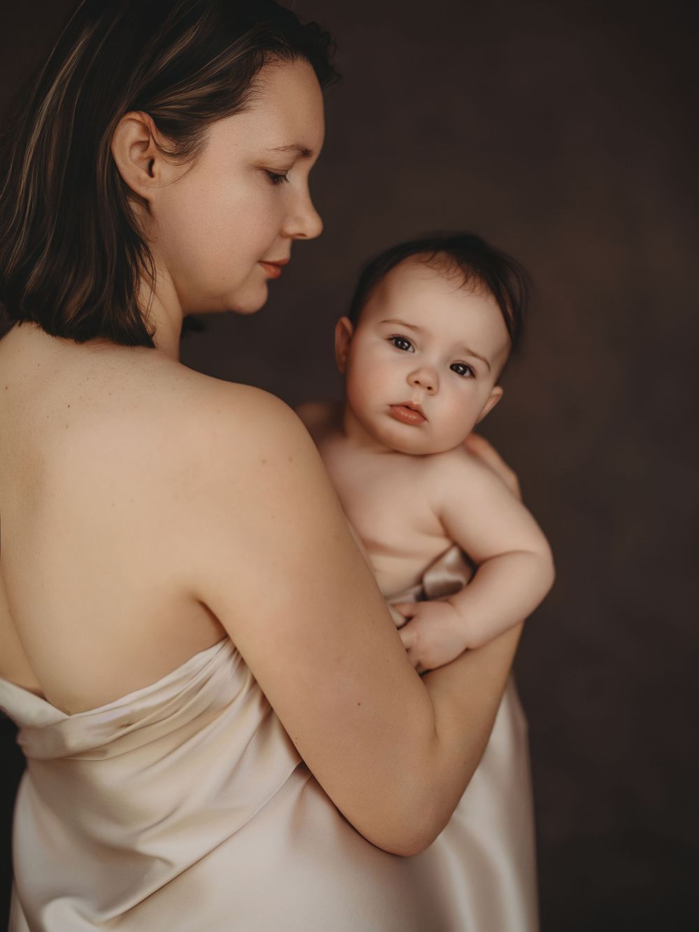 A mother in a white silk dress tenderly holds her baby against a dark studio background.