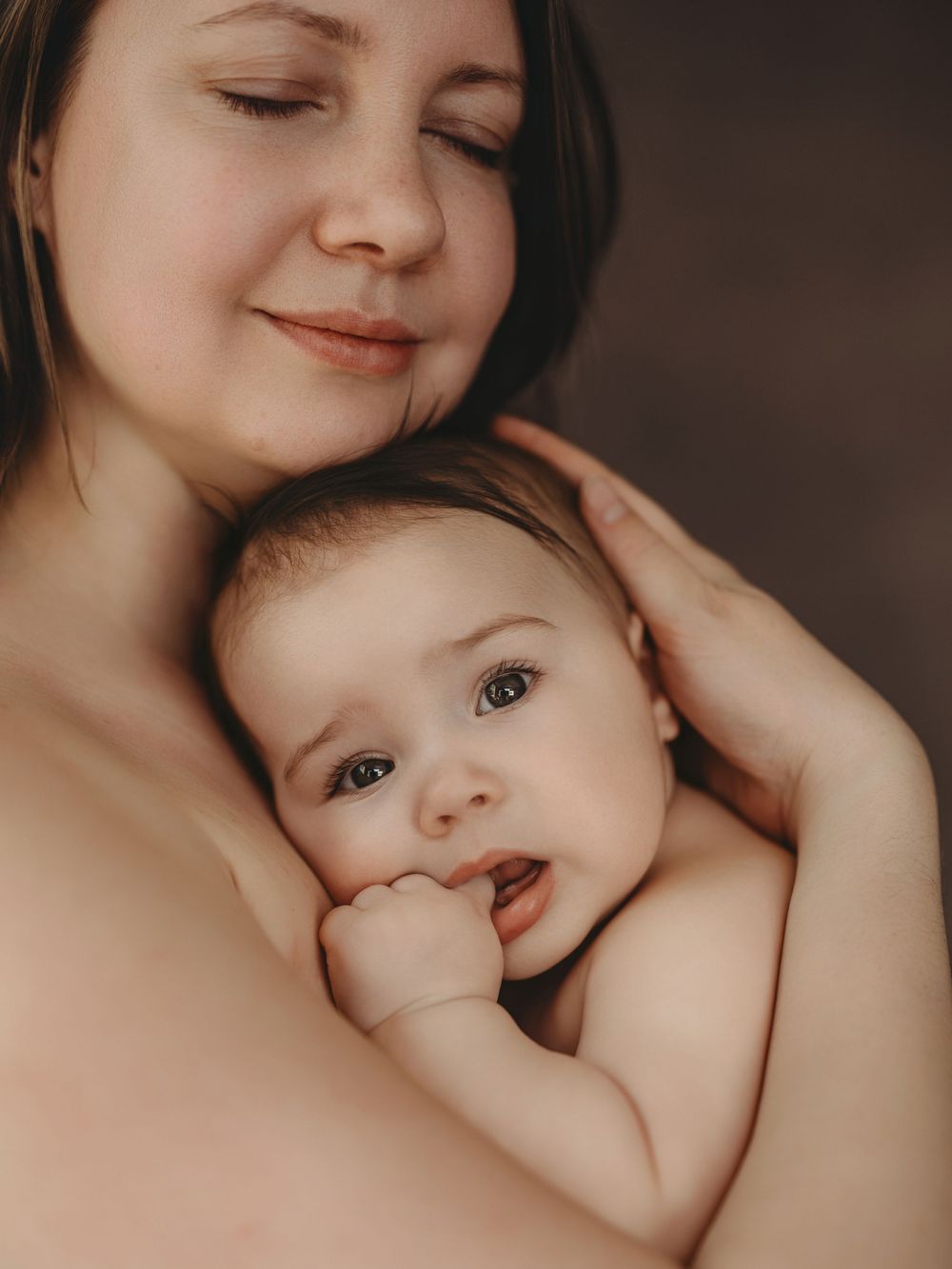 A mother tenderly holds her baby against her chest, smiling softly with closed eyes while the infant gazes at the camera.