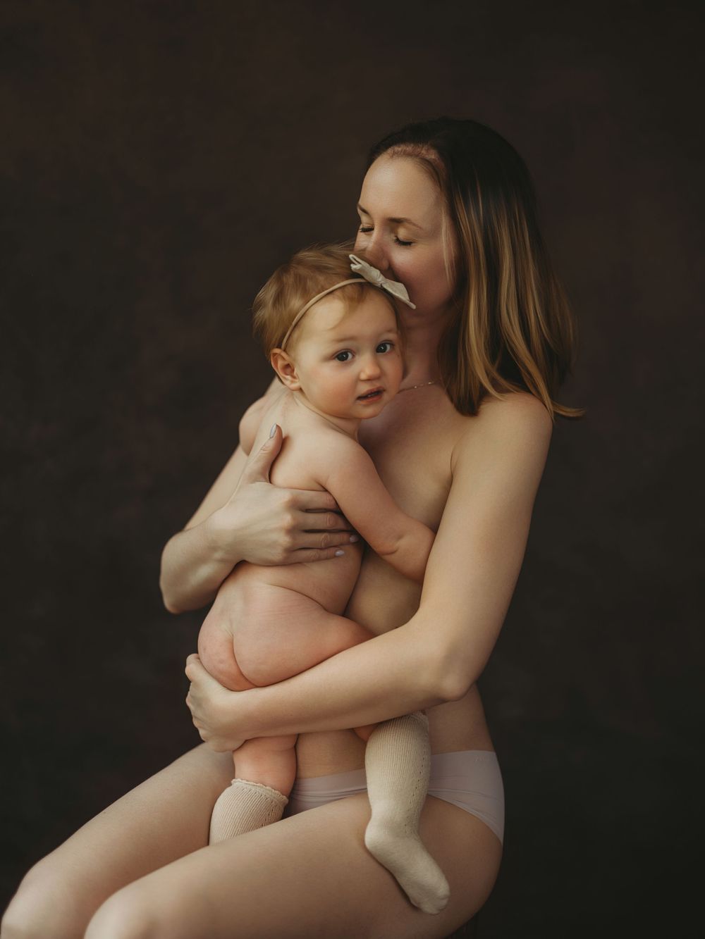 A mother tenderly holds her baby in a warm studio portrait against a dark background.