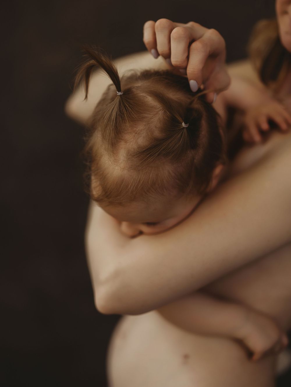 A toddler girl with pigtails rests her head on an adult's shoulder in a tender embrace.