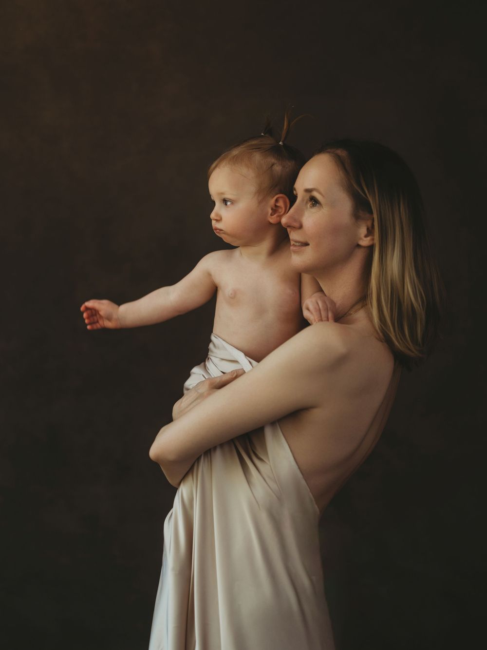 A mother tenderly holds her baby in a soft, artistic studio portrait with warm, dark background lighting.