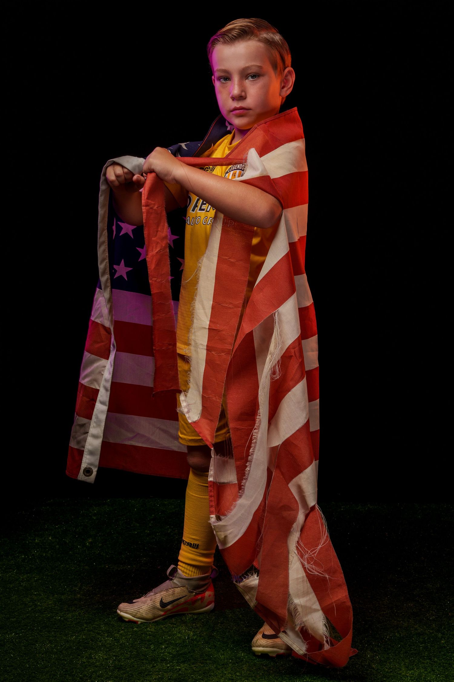 Child draped in American flag standing on grass against dark background.