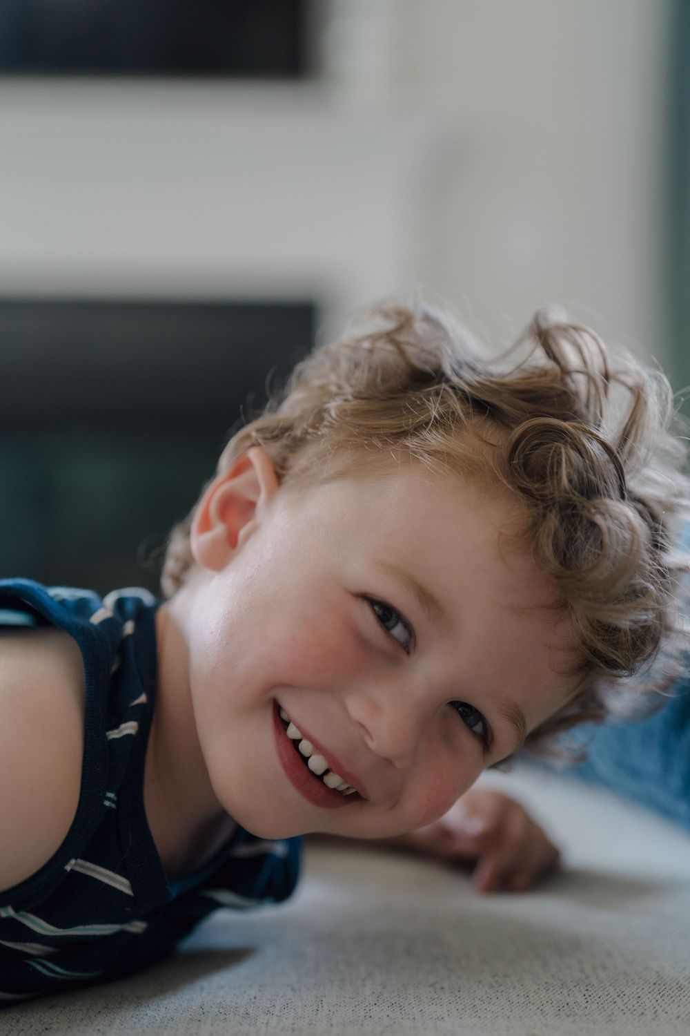 A joyful young child with wavy blonde hair lies on the floor while wearing a blue striped shirt with a bright smile at in home Kent, OH pic
