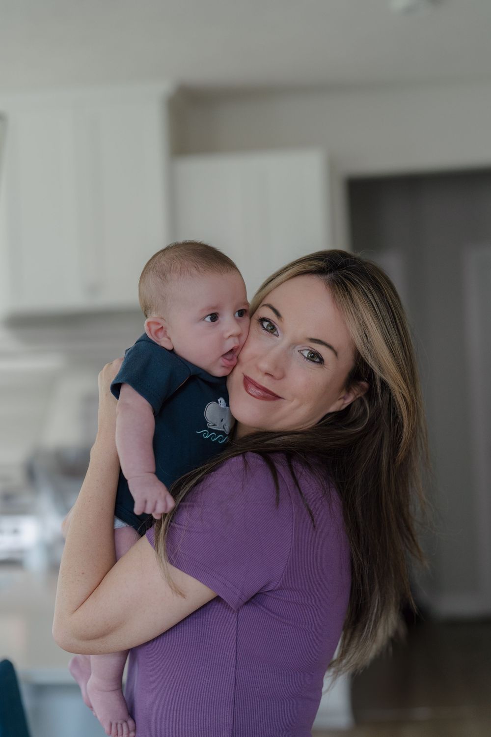 A baby in a navy onesie is held by someone wearing a purple shirt in a bright kitchen setting at in-home newborn photo session in Kent, Ohio
