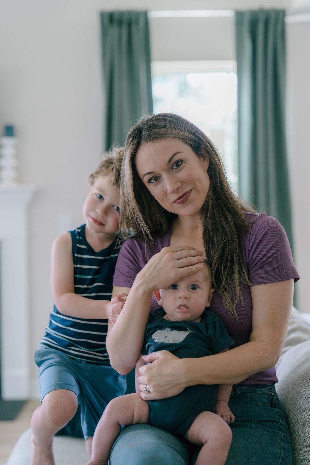 A young woman in a purple shirt sits with two children on a couch in front of turquoise curtains in a bright room at newborn photo session