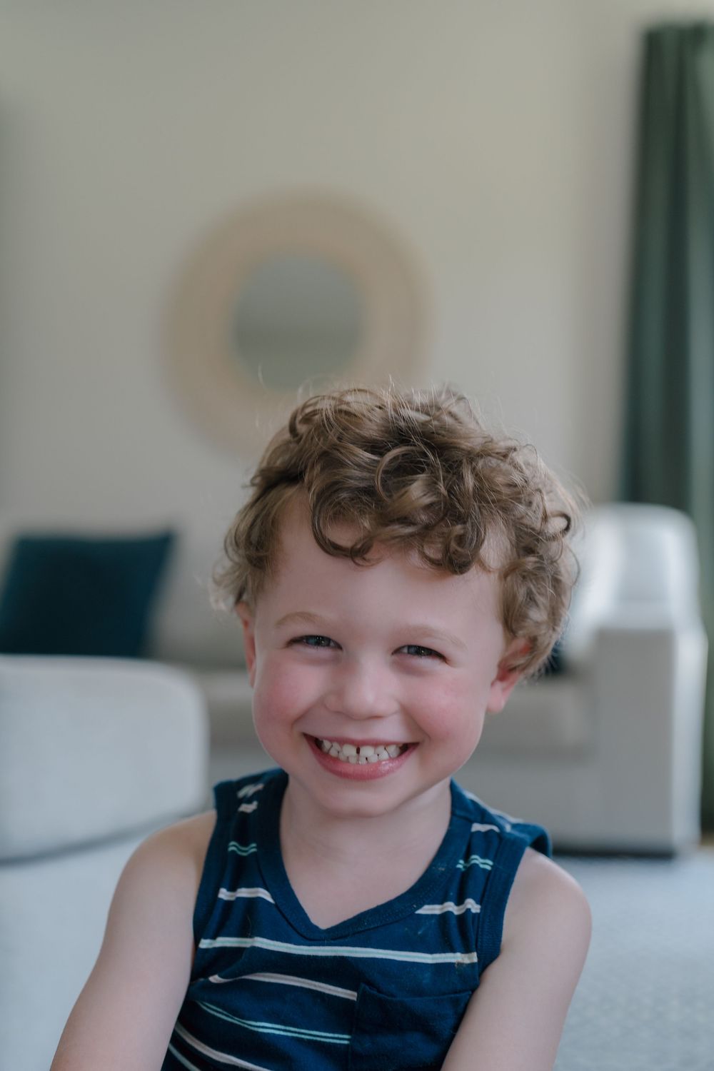 Young child wearing a blue striped tank top sits and smiles indoors with white walls and decor visible in background of Kent, Ohio session