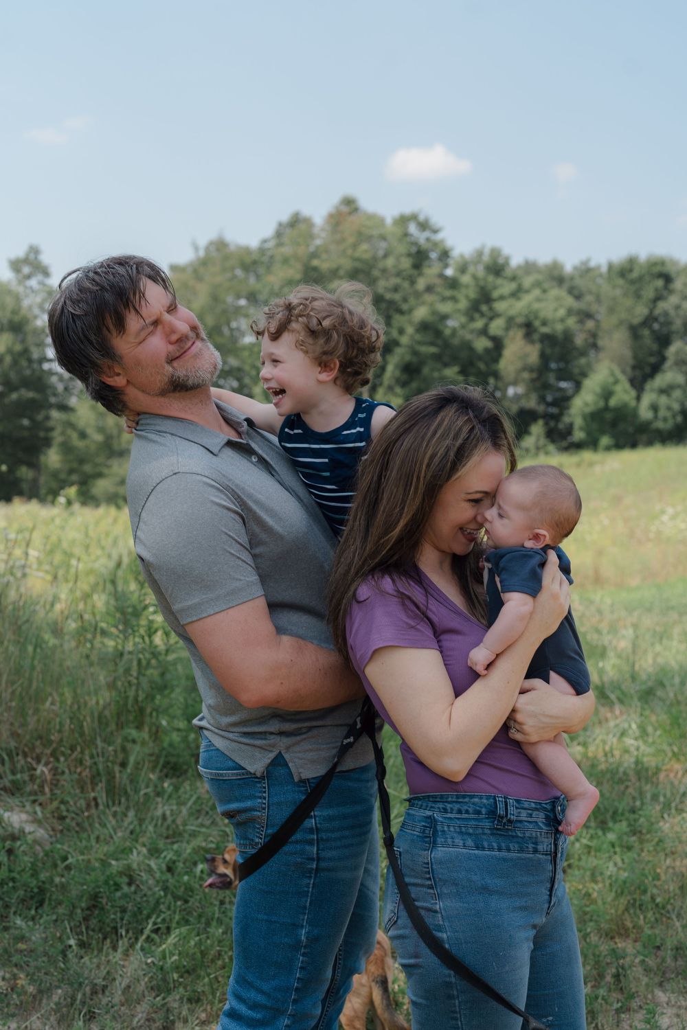 A family shares joyful moments together while playing and embracing in a sunny meadow with green trees behind at newborn photo session in OH