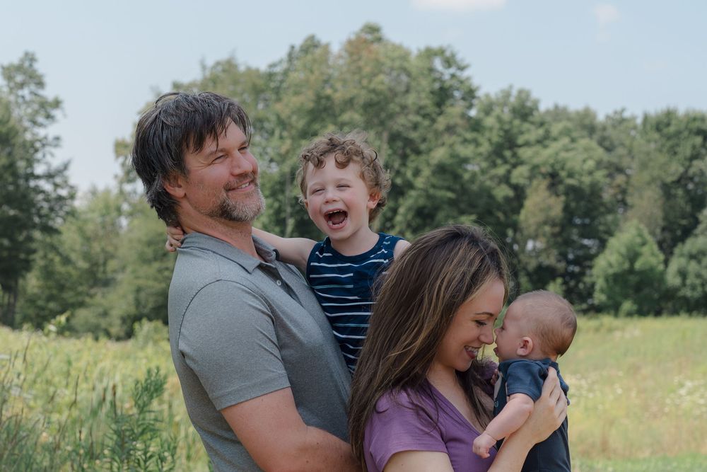Happy family enjoying a playful moment together in a sunny meadow with green trees in the background of newborn photo session in Kent, Ohio