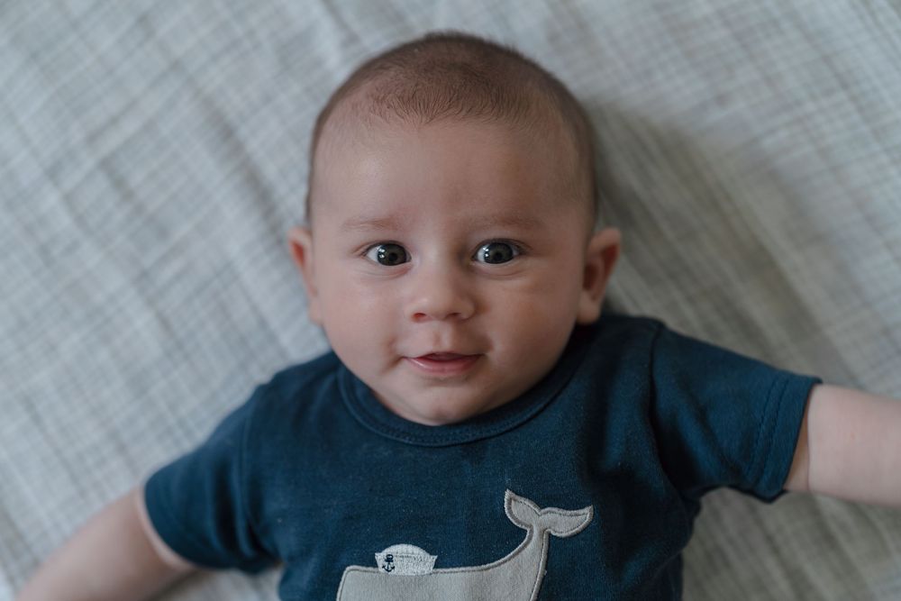 Baby wearing navy blue onesie smiles while lying on gray fabric bedding at Kent, Ohio Newborn Photo session