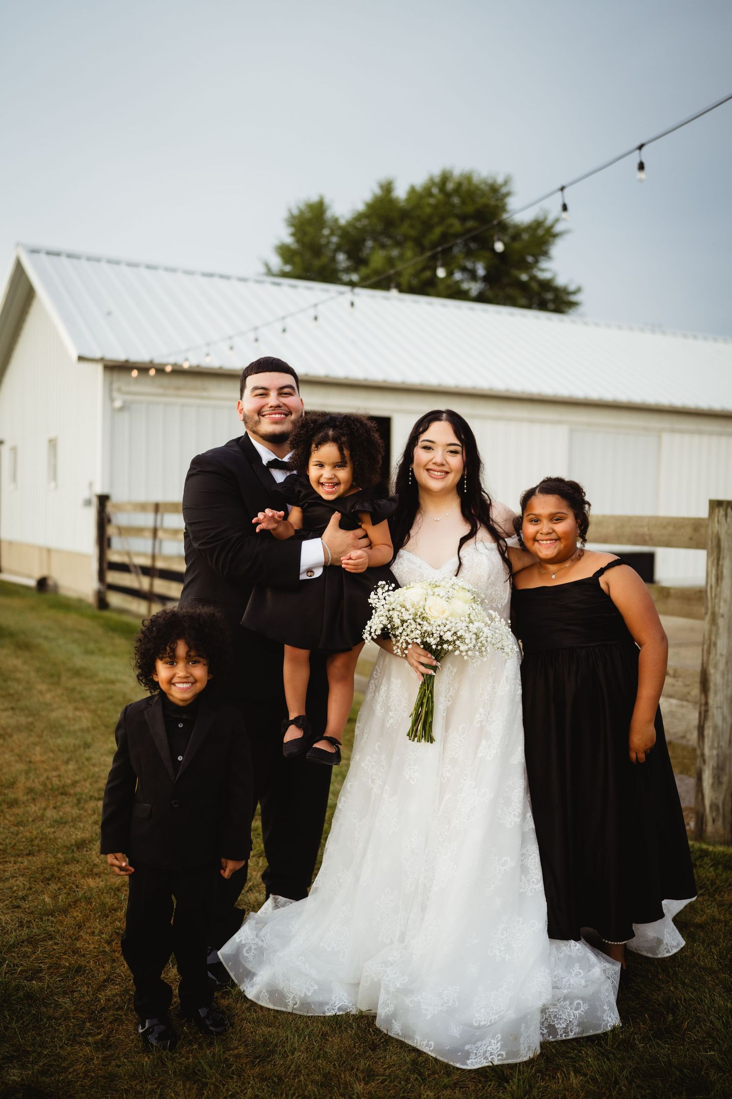 A group poses together in formal attire outside a white barn building during an outdoor wedding celebration.