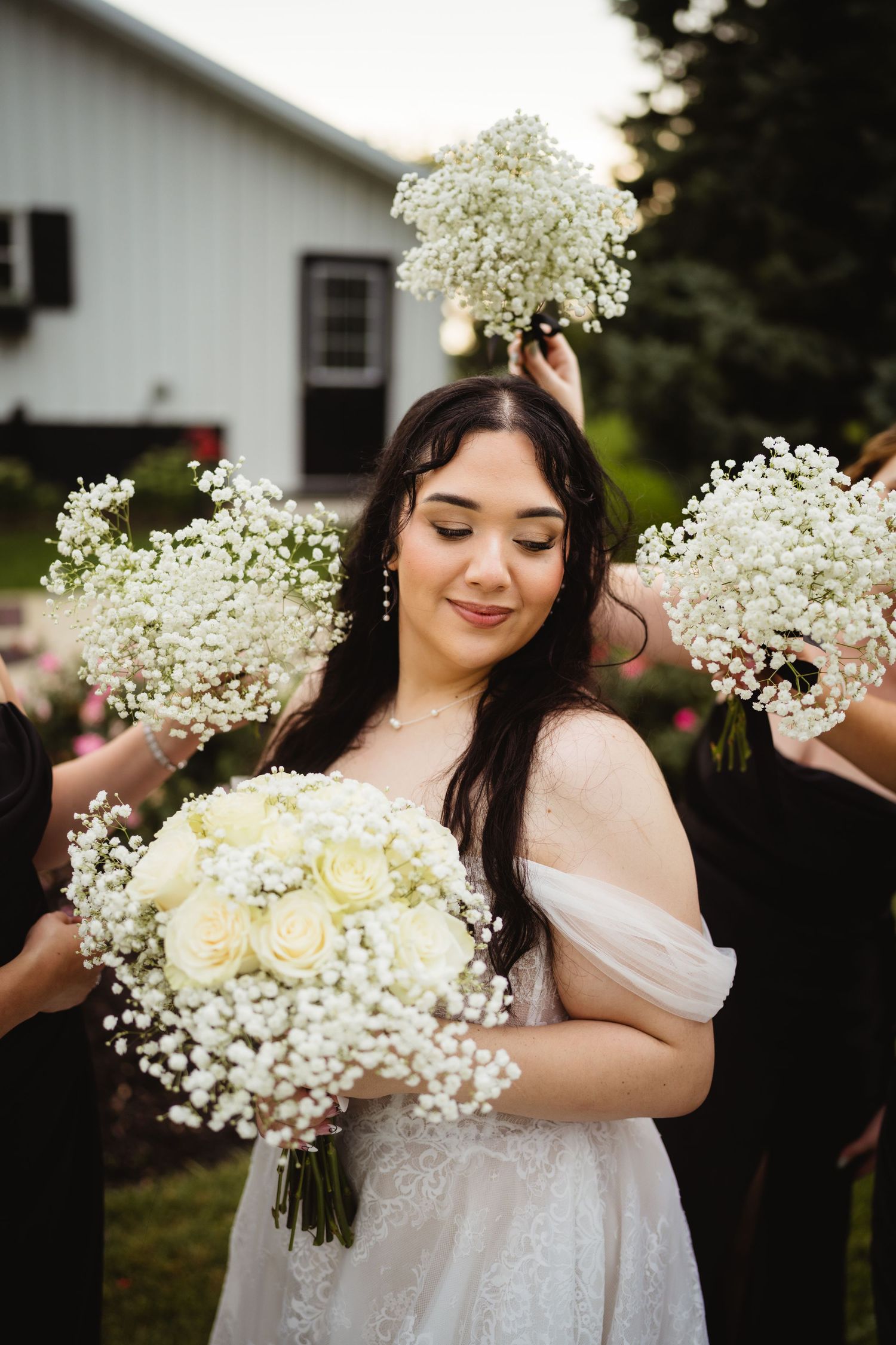 A bride in a flowing white dress holds a bouquet of white roses and baby's breath while standing outdoors near a house.
