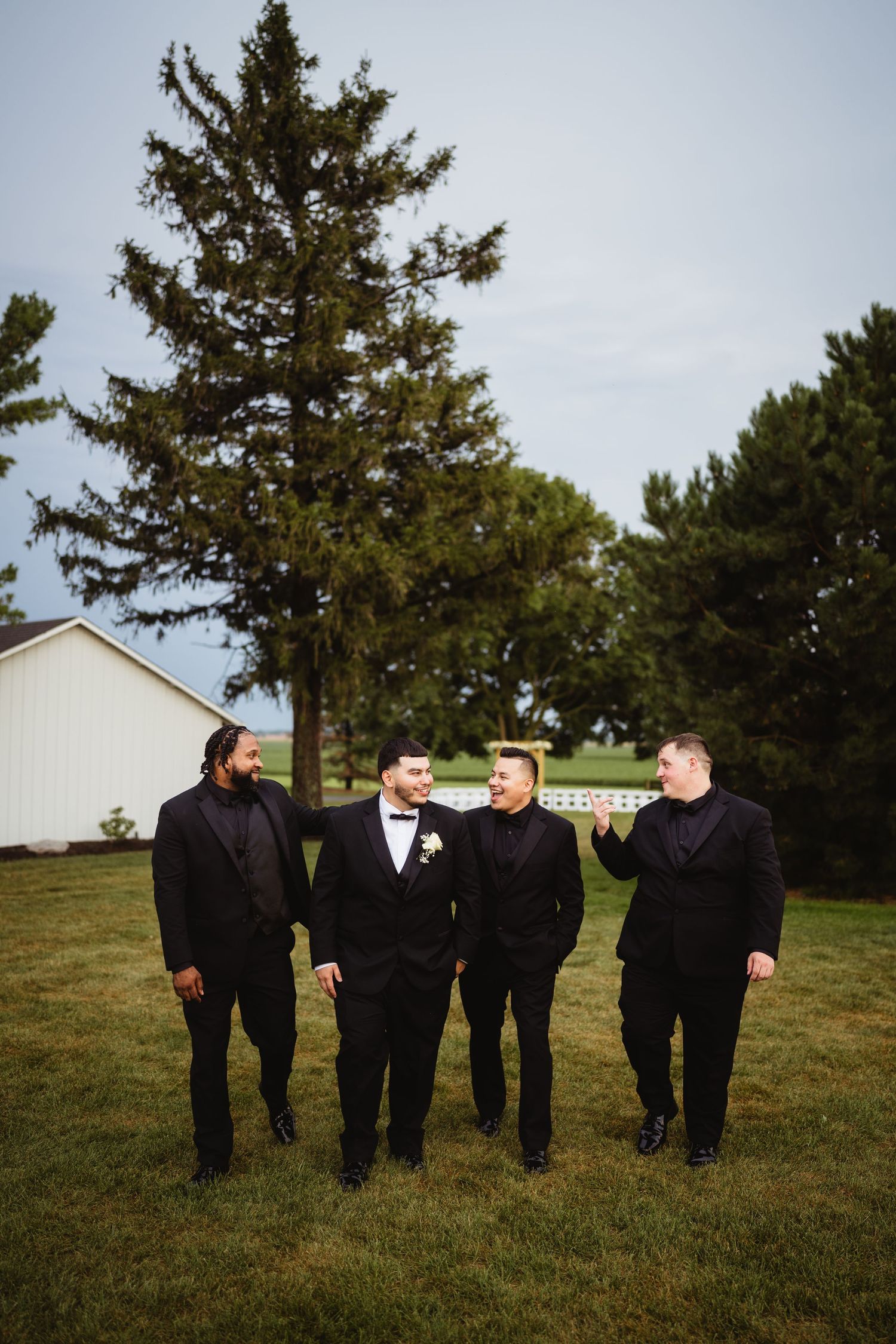 A group in formal black attire stands together on a grassy lawn with evergreen trees and a white building in the background.