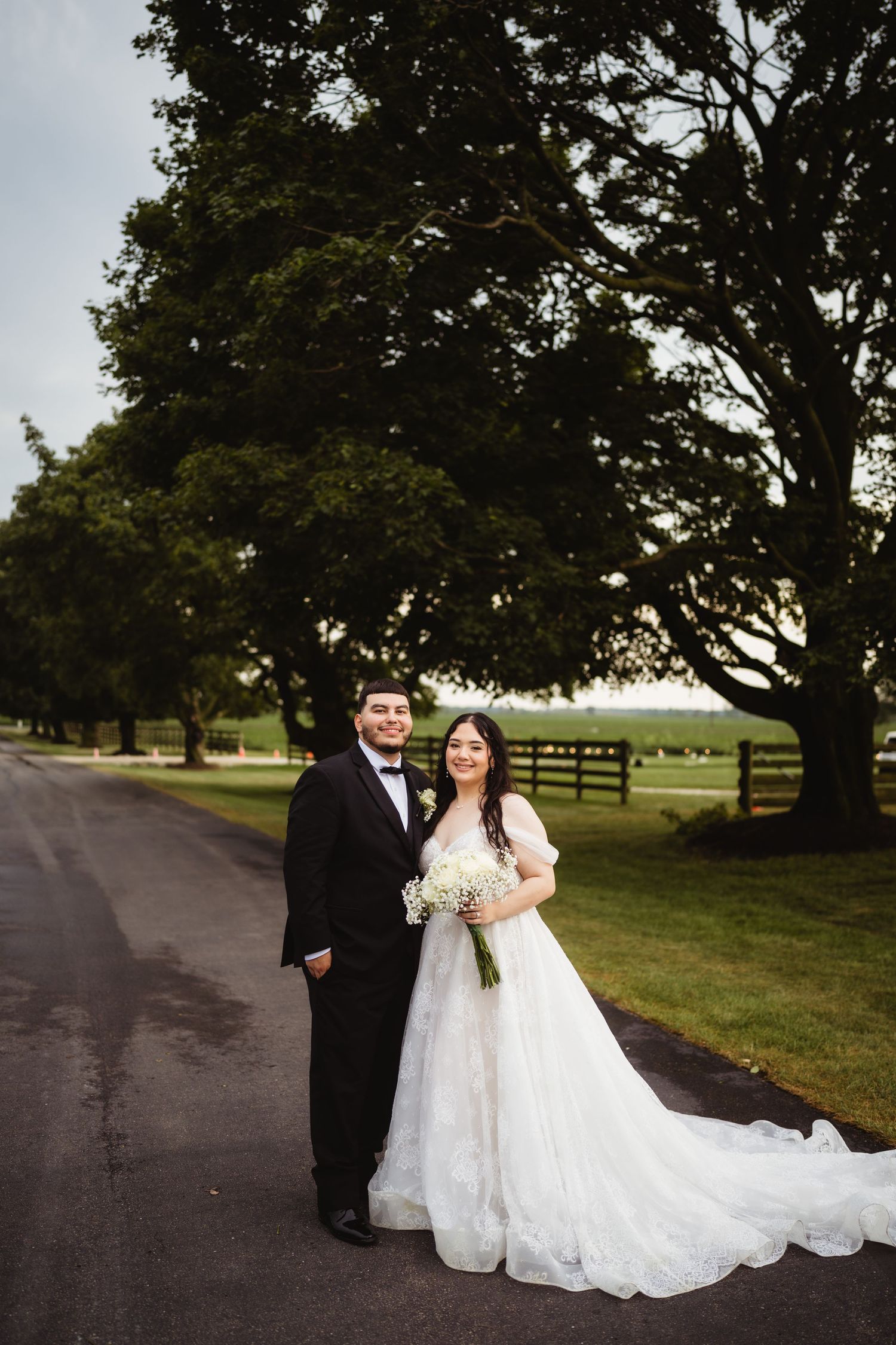 A wedding couple stands on a country road with large trees and wooden fences creating a romantic rural backdrop.