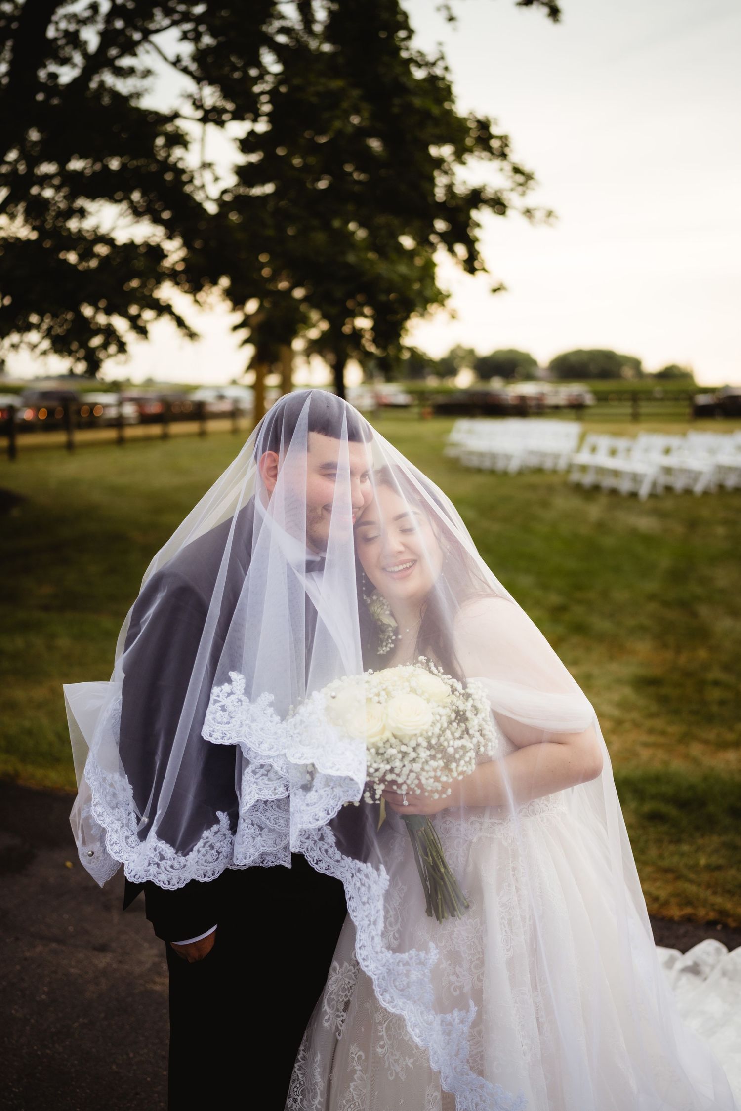A couple shares a romantic moment under a white lace veil at an outdoor wedding ceremony with folding chairs visible.