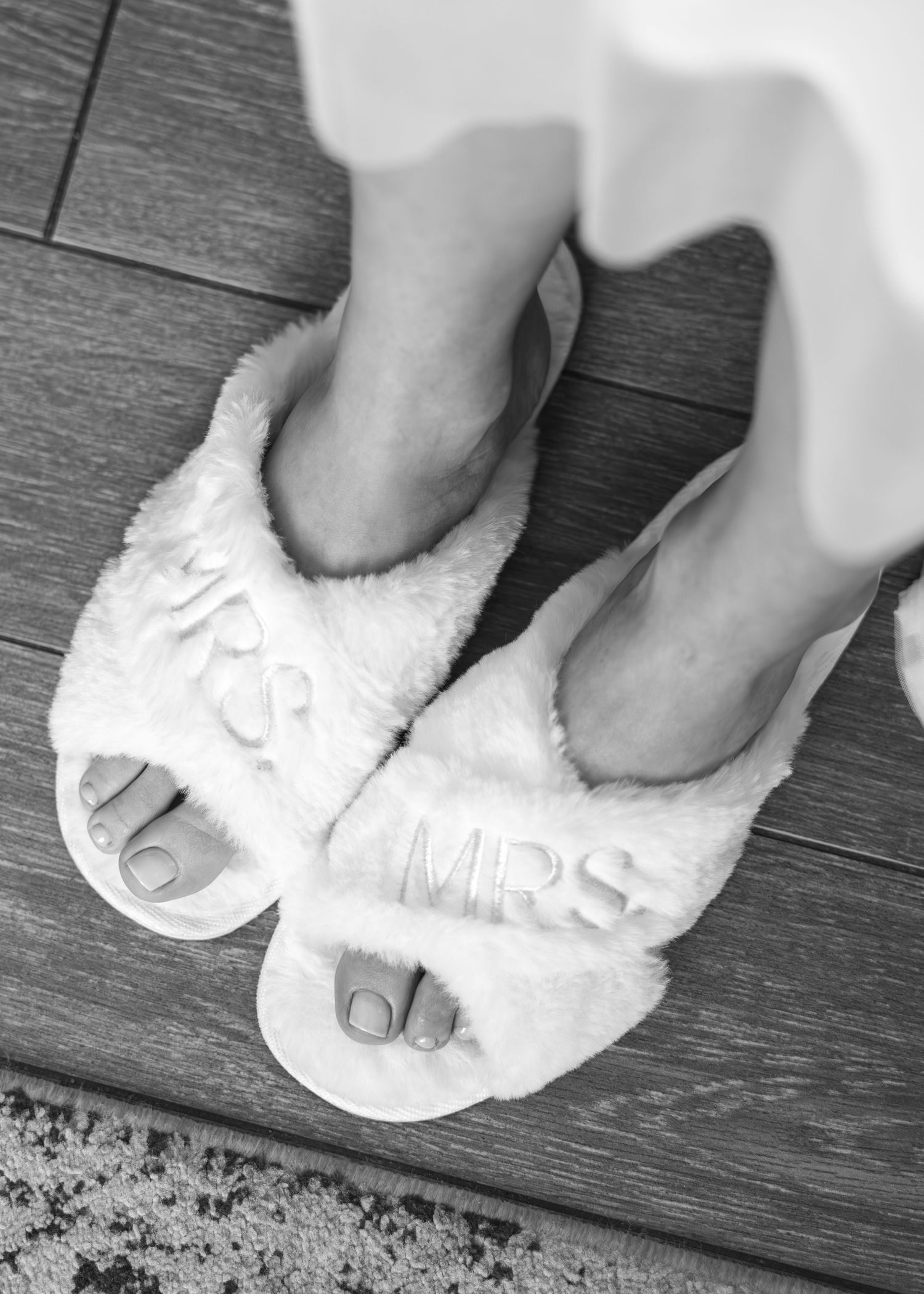 A pair of plush white MRS slippers on a dark wooden floor, showing freshly pedicured feet.