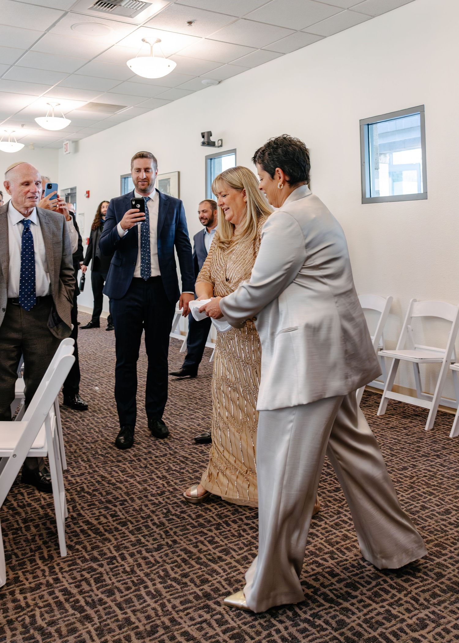 Wedding guests in formal attire gather and chat in a carpeted hallway.