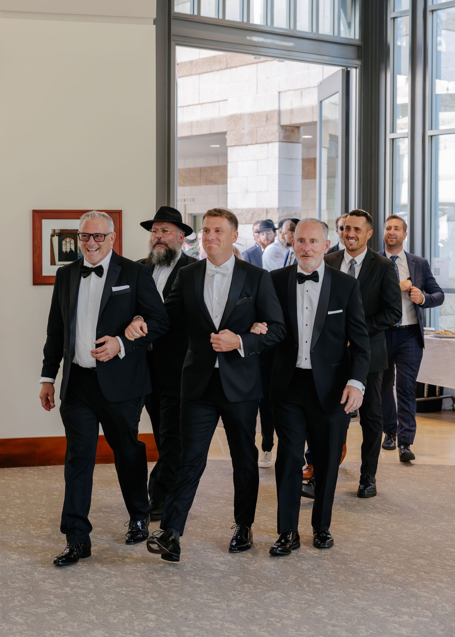A group of groomsmen in black tuxedos and bow ties walk together down a hallway.