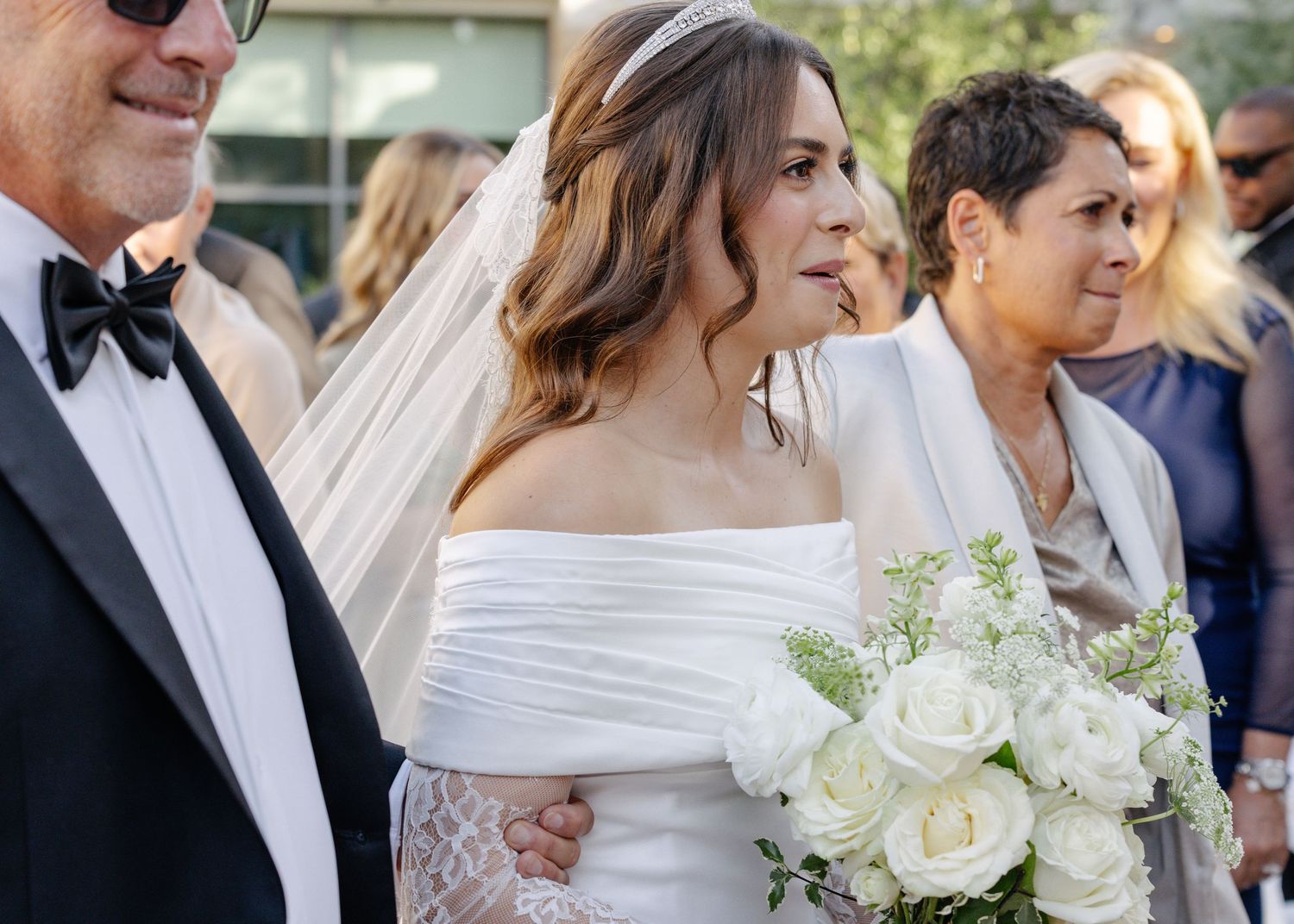 A bride in an elegant off-shoulder white gown holds a classic white rose and greenery bouquet at an outdoor wedding ceremony.