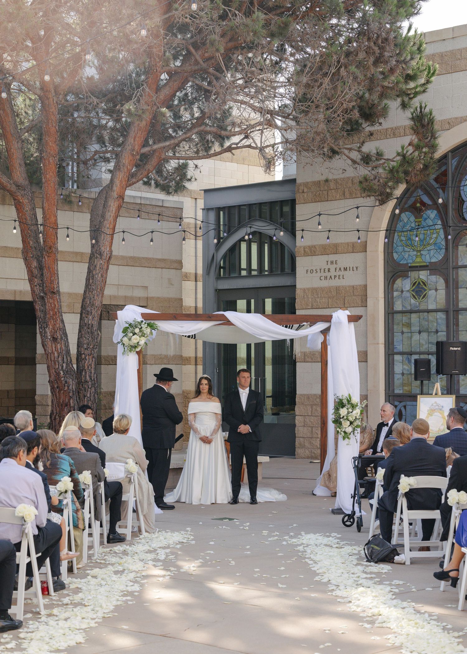 Romantic outdoor wedding venue setup with white umbrellas, string lights, and scattered flower petals along a natural pathway.
