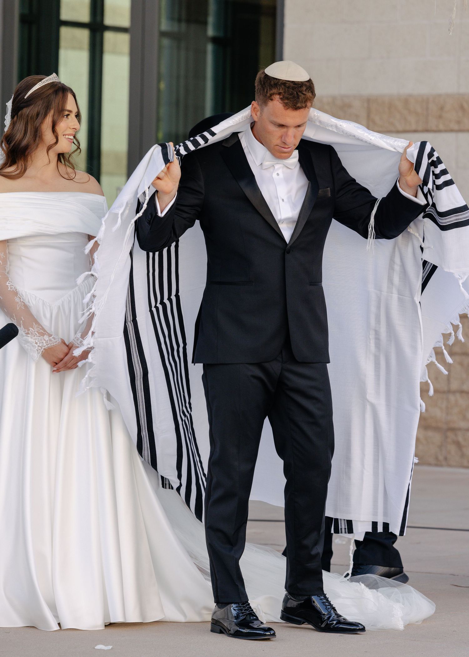 A couple at their wedding with the groom holding up a traditional prayer shawl while bride wears elegant white dress.