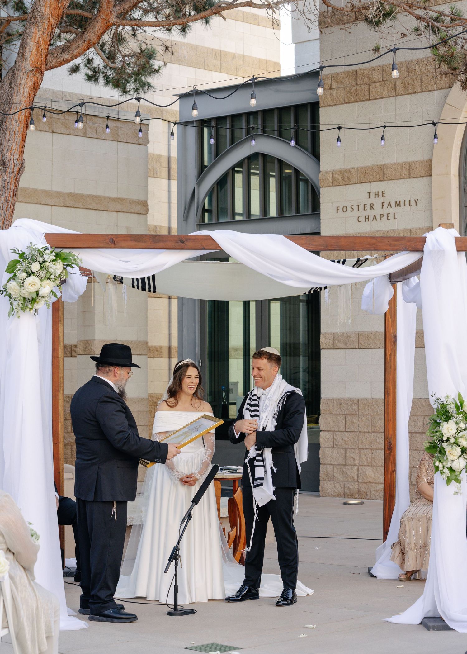 Wedding ceremony setup with white drapery and floral arrangements at a stone building venue with string lights overhead.