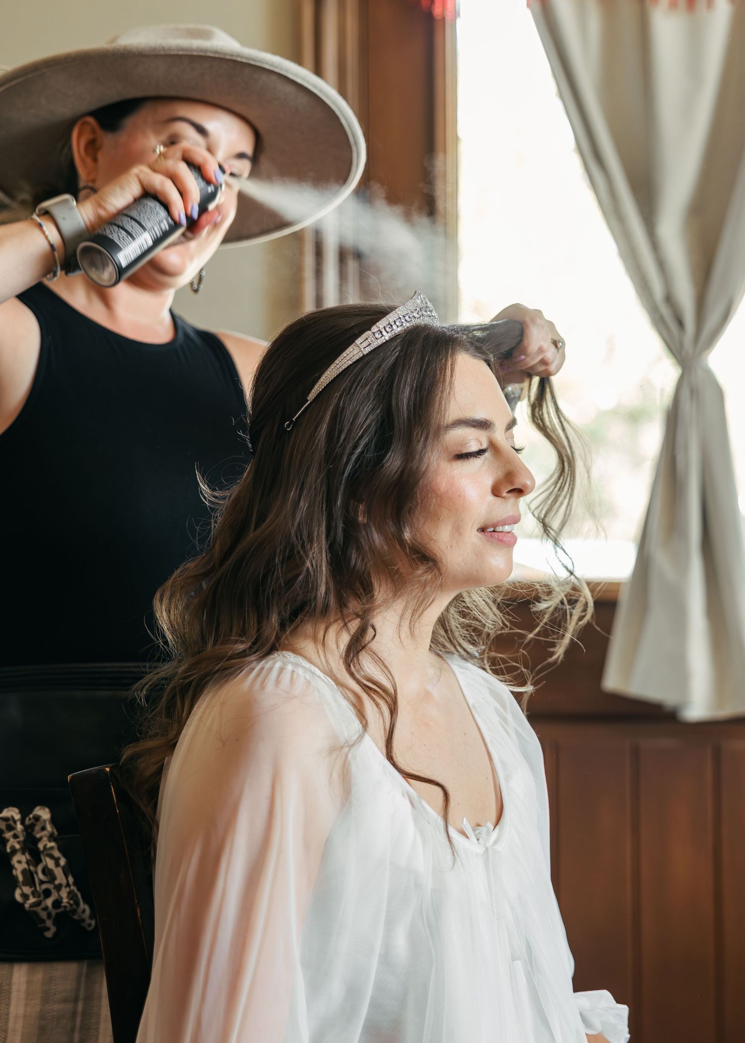 Hairstylist uses hairspray while styling client's wavy brown hair in a sunlit room with sheer curtains.