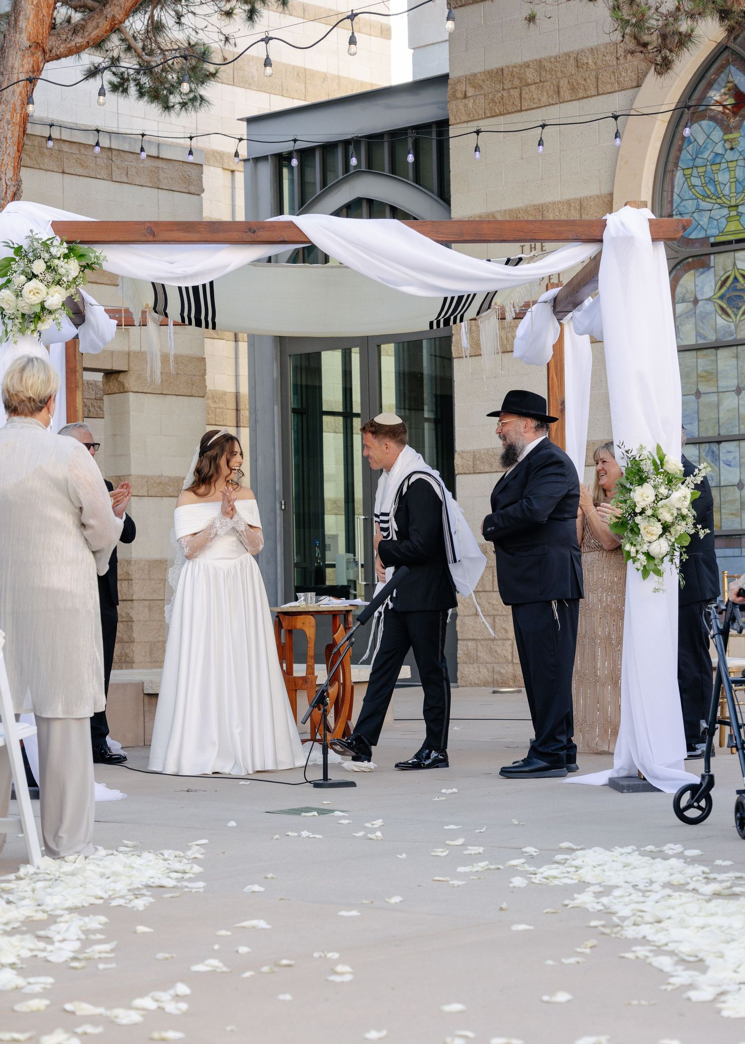 Elegant wedding ceremony taking place outdoors against stone building walls with white rose petals scattered on ground.
