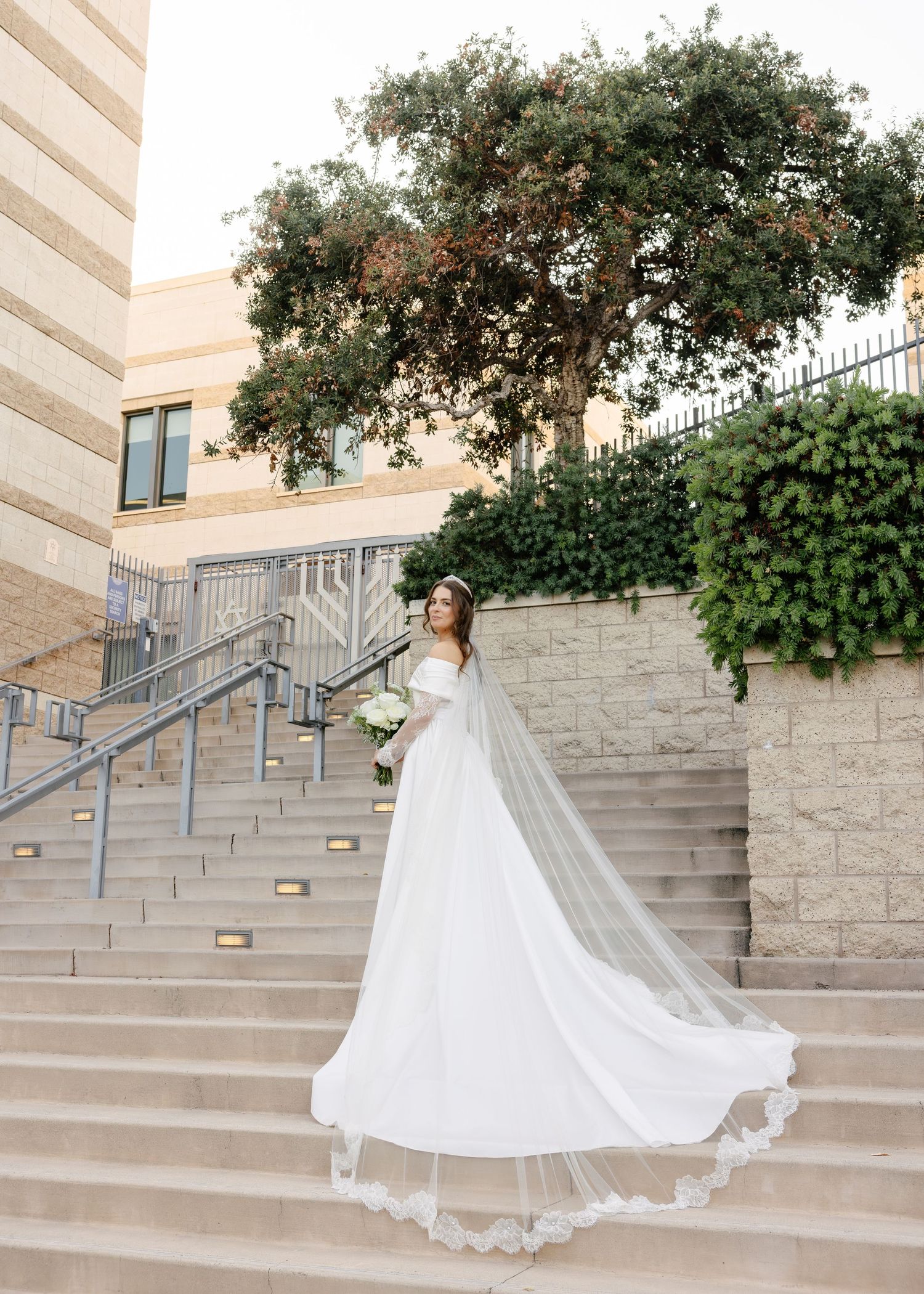 An elegant wedding dress with flowing train cascades down stone steps against a modern building backdrop and greenery.