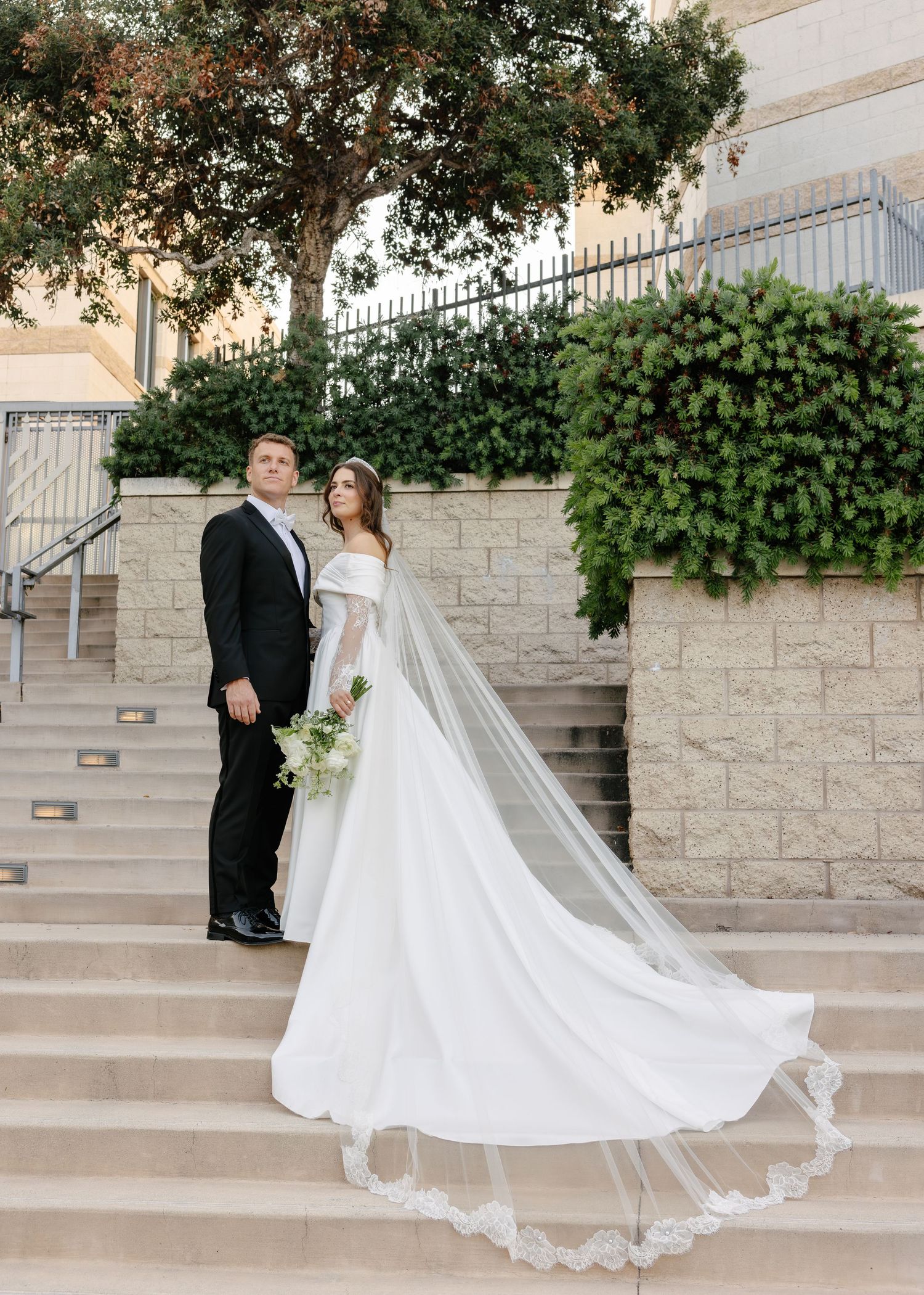 A wedding couple stands on grand stone steps with their flowing dress train creating a dramatic scene.