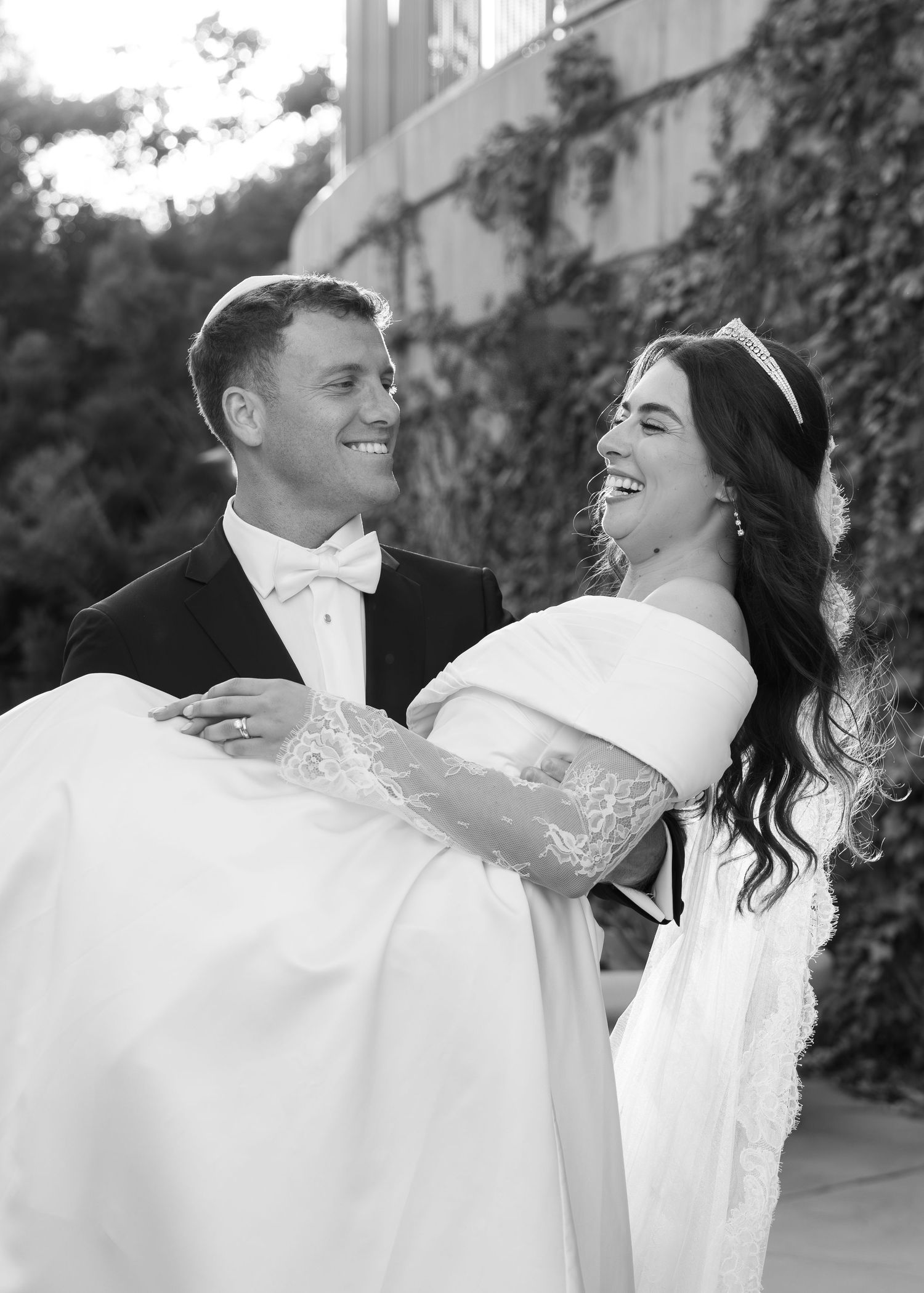 Black and white wedding photo of a couple sharing a joyful moment as the groom carries his bride in a traditional pose.