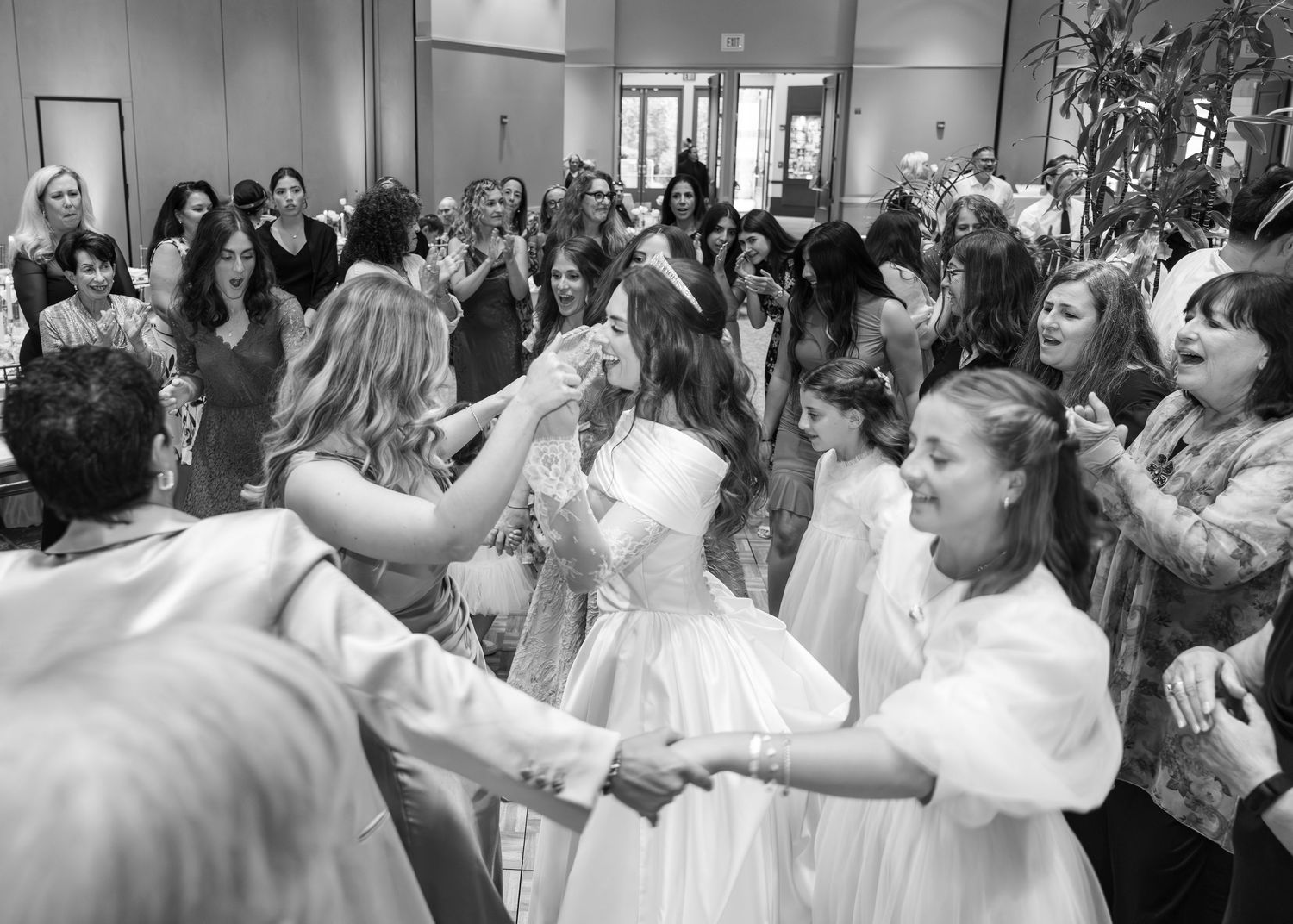 People dancing together in a crowded ballroom during a celebration while wearing white and light colored formal attire.