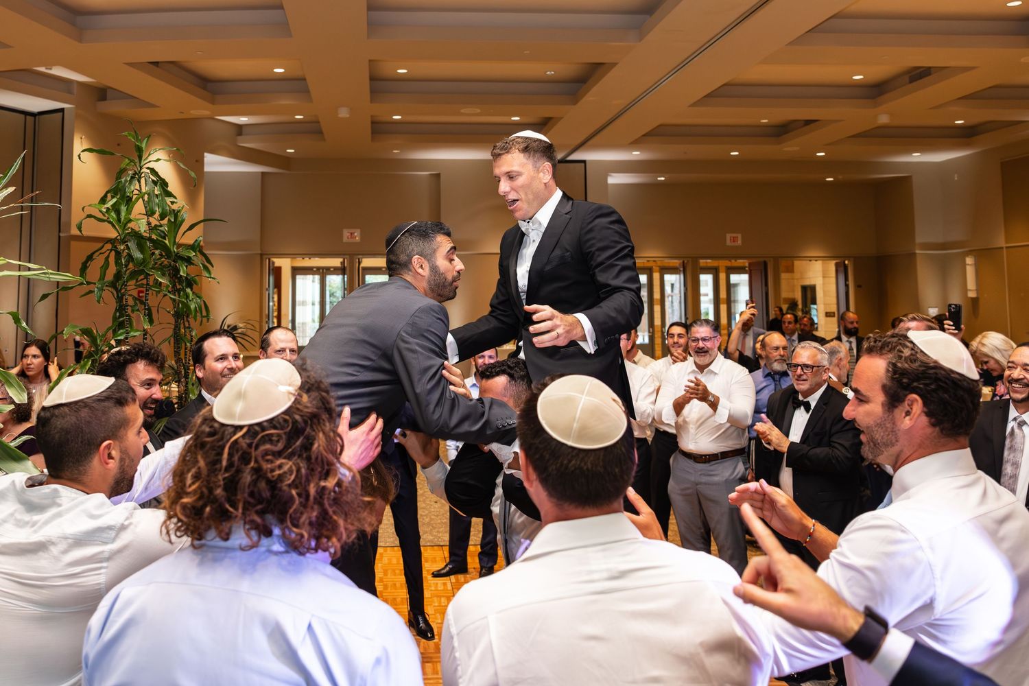Orthodox Jewish men joyfully dance in a circle during a celebration at an indoor venue.