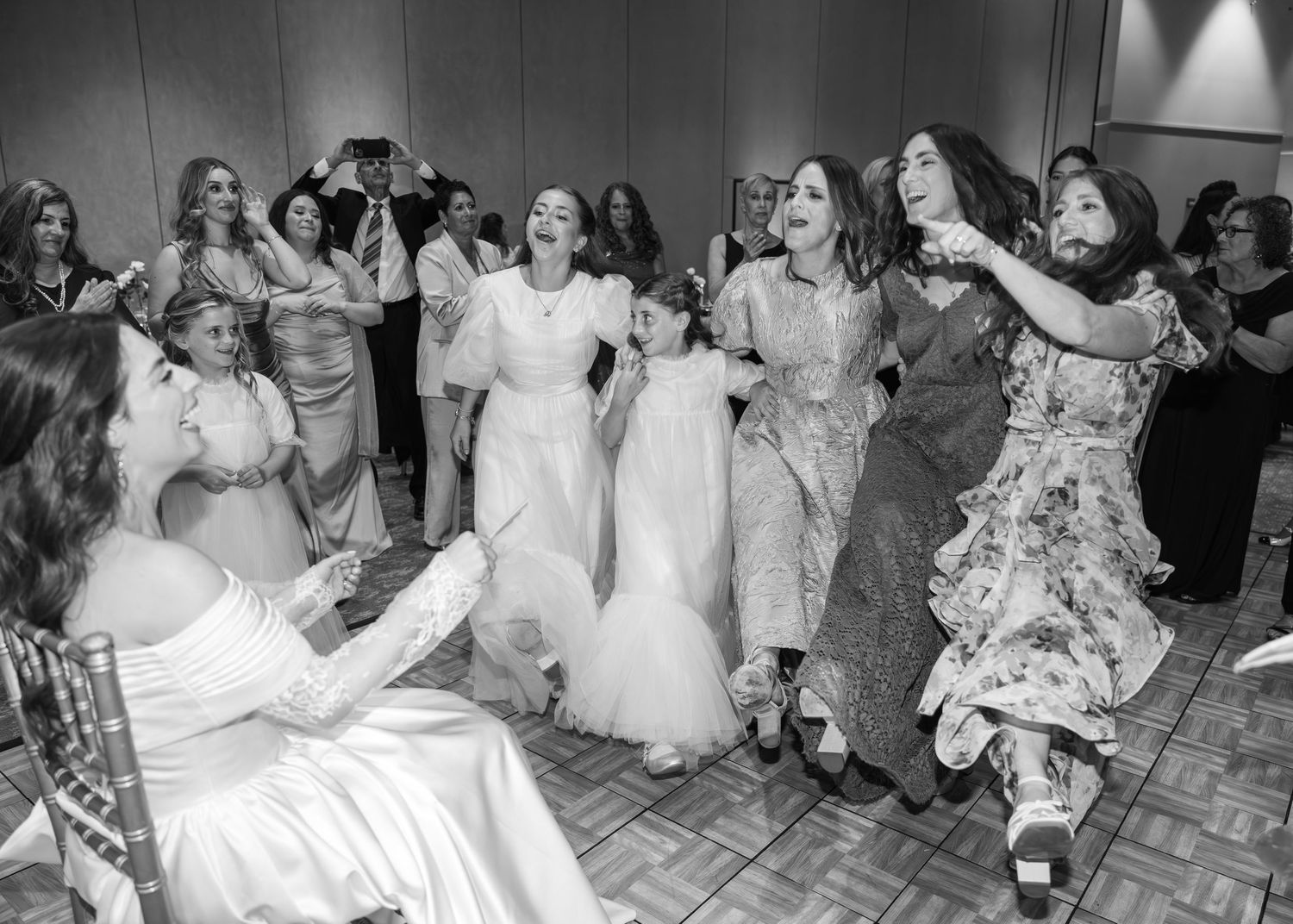 A joyful group dances together at a wedding reception in black and white photography.