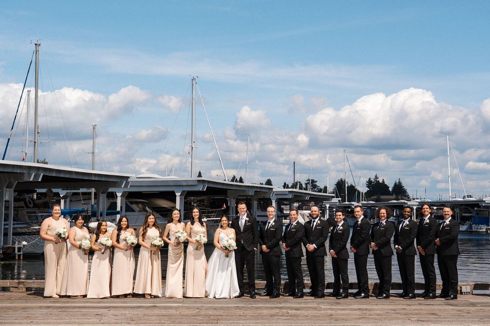 Wedding party in formal attire lined up along a waterfront dock with boats and blue sky in the background.