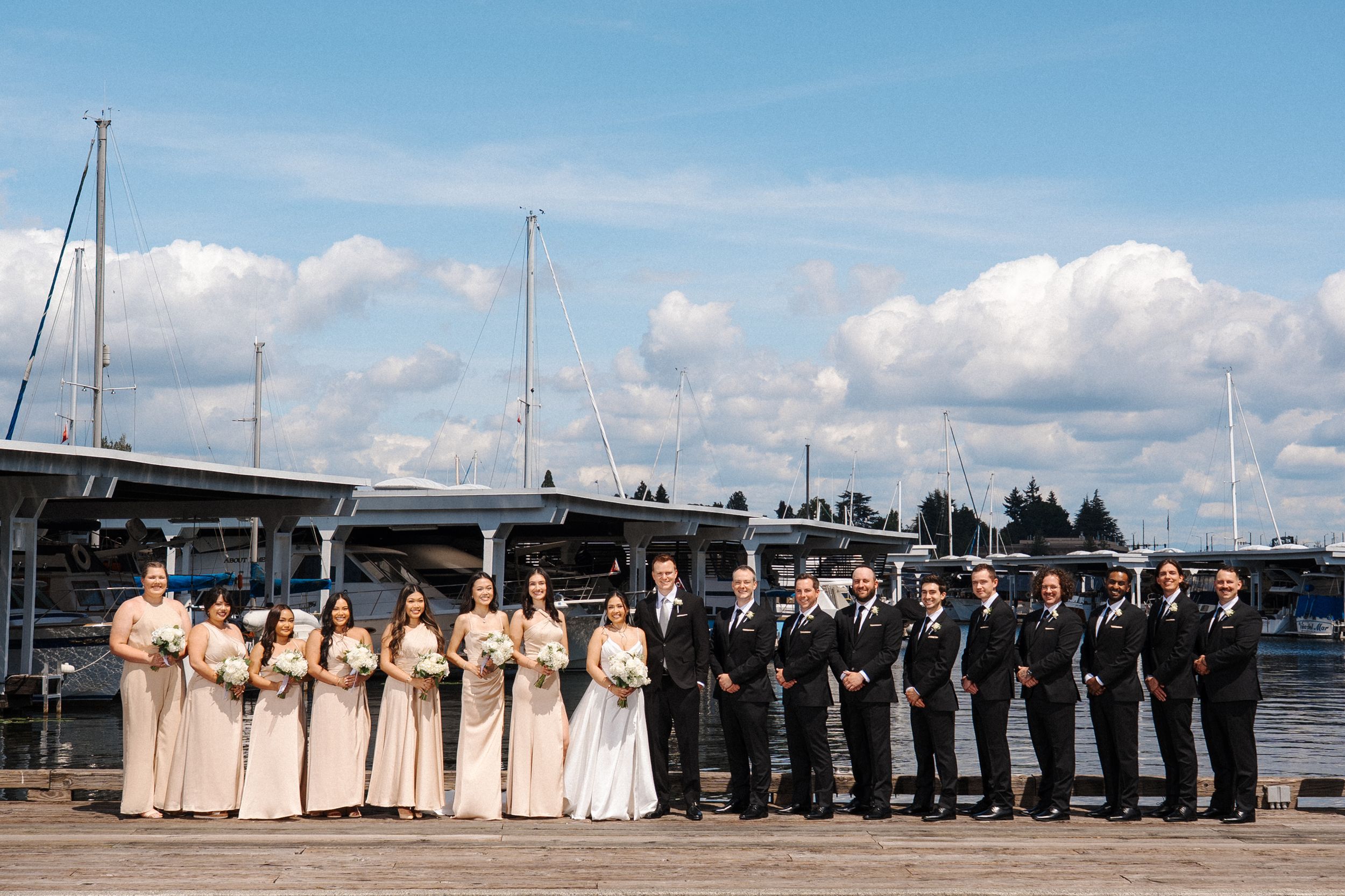 Wedding party in formal attire lined up along a waterfront dock with boats and blue sky in the background.