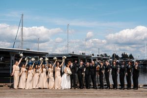 Wedding party lined up on dock with bridesmaids in beige dresses and groomsmen in dark suits by water.