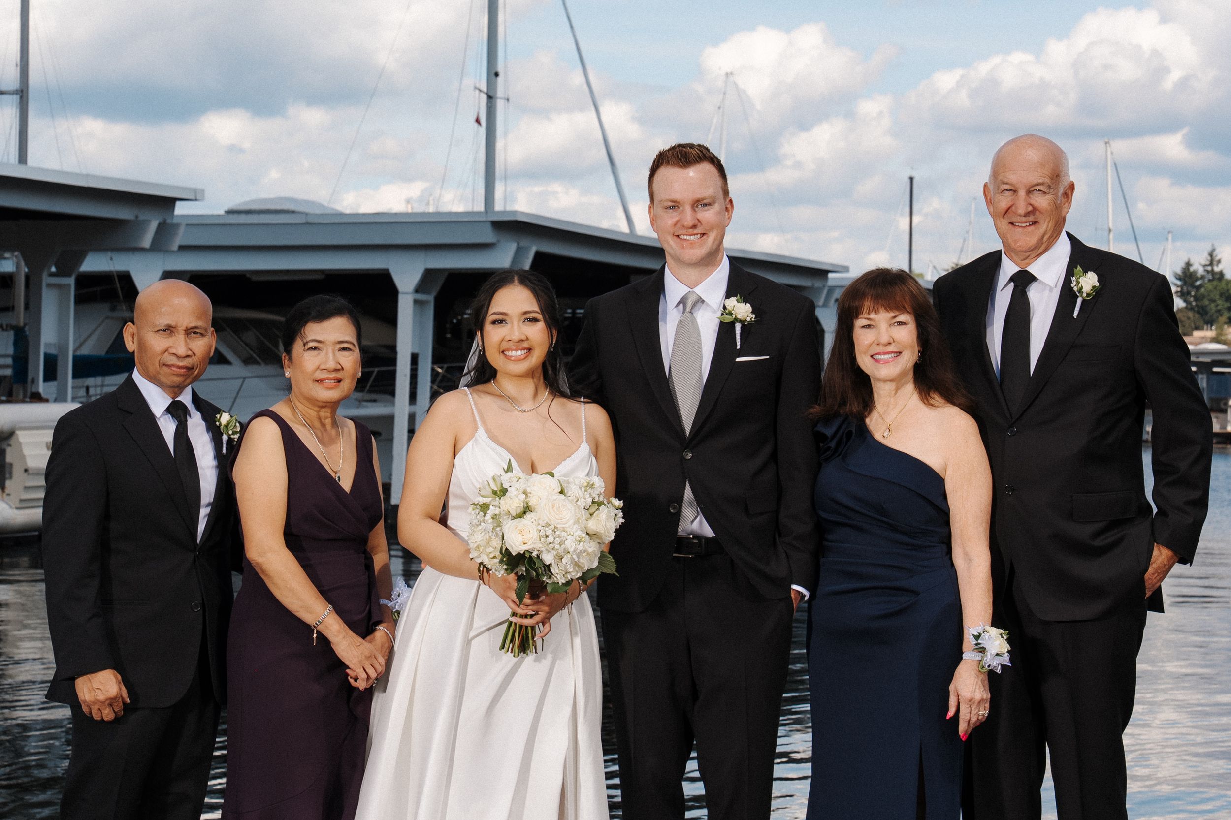 Wedding party in formal attire poses near water with boats and dock in background.