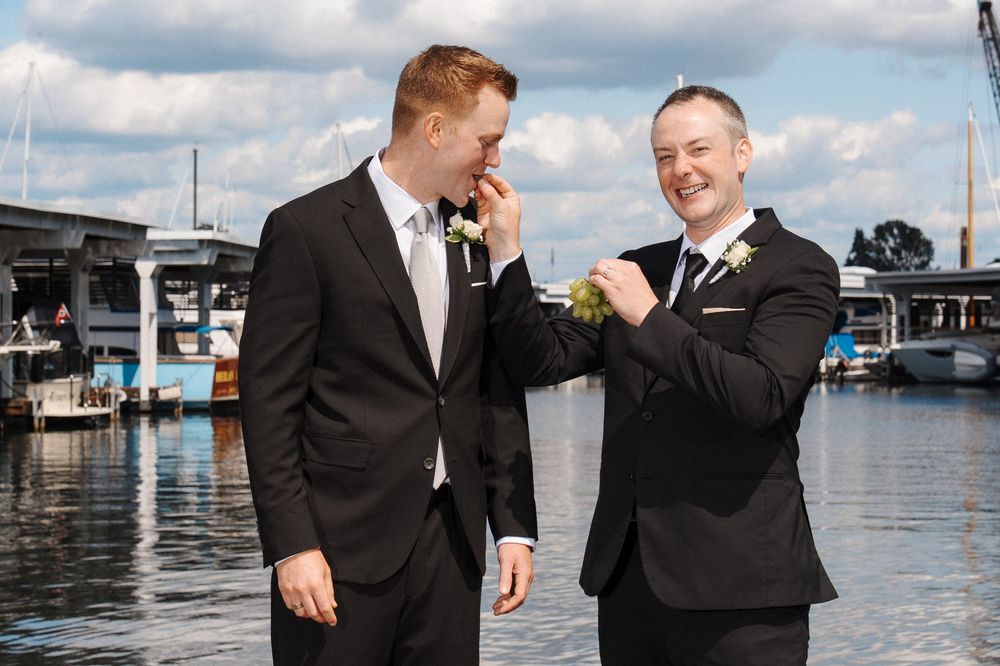 Wedding party members in black suits laugh together on a dock by the water on a sunny day.