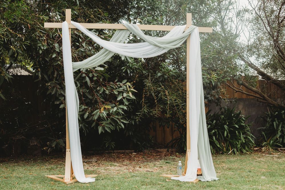 Elegant white fabric drapery and floral decor create a romantic outdoor wedding ceremony arch in a garden setting.