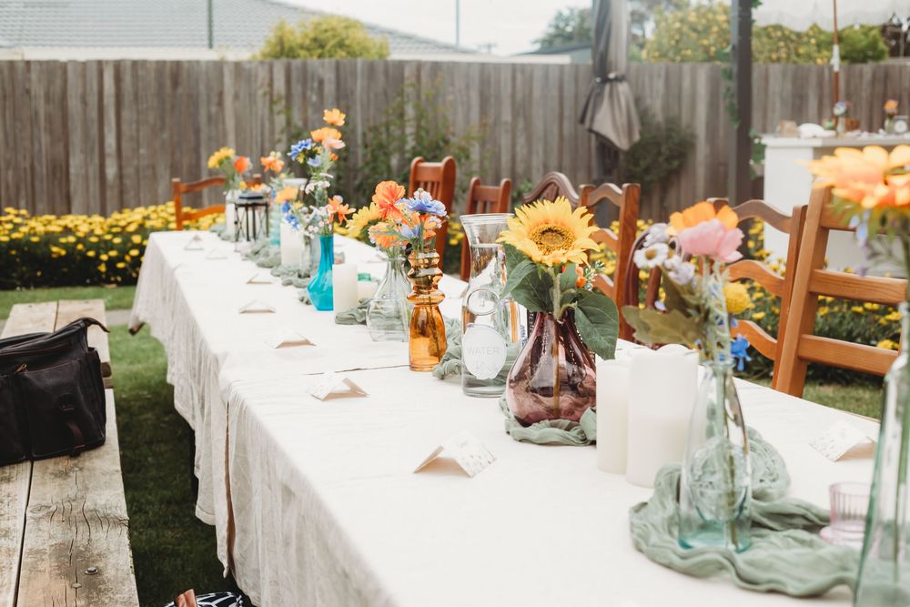 A long white table decorated with colorful floral centerpieces, glass vases and wooden chairs for an outdoor garden party.
