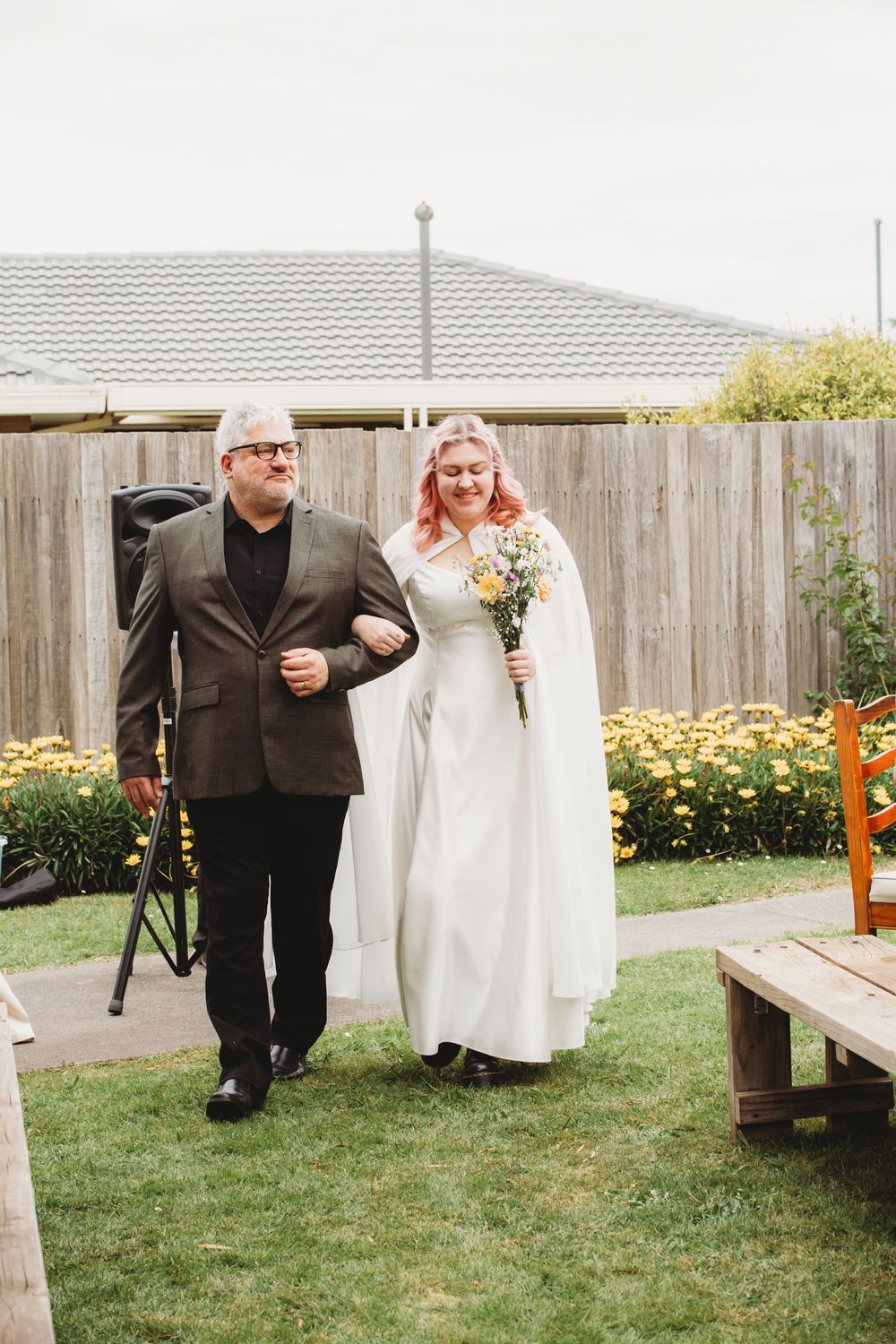 Walking down the garden aisle together on a beautiful outdoor wedding day with yellow flowers in the background.