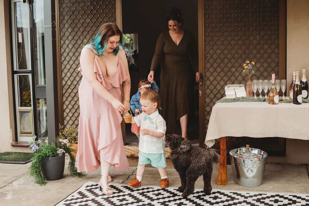 A baby in mint green clothing reaches out while standing between two people wearing pink and black outfits.