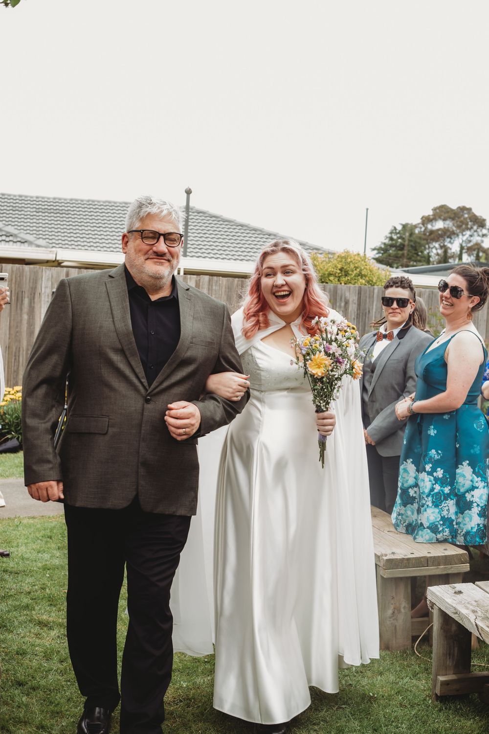 Wedding party gathered outdoors in formal attire with white and teal dresses against a garden backdrop.