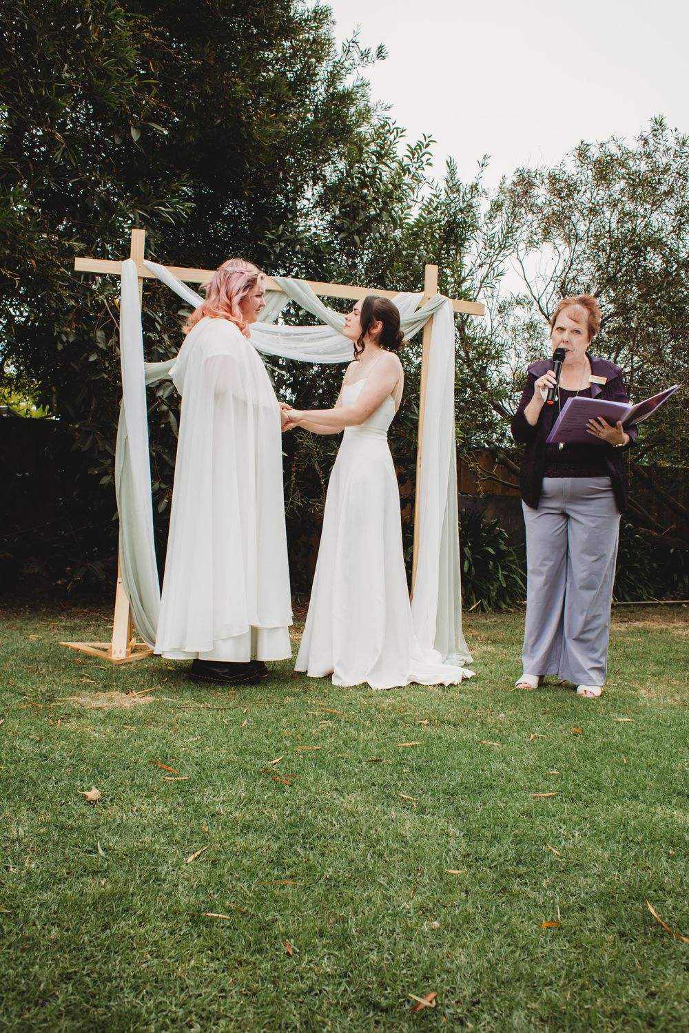 Four people in wedding attire gather around a white arbor draped with fabric on a grassy lawn during a ceremony set-up.