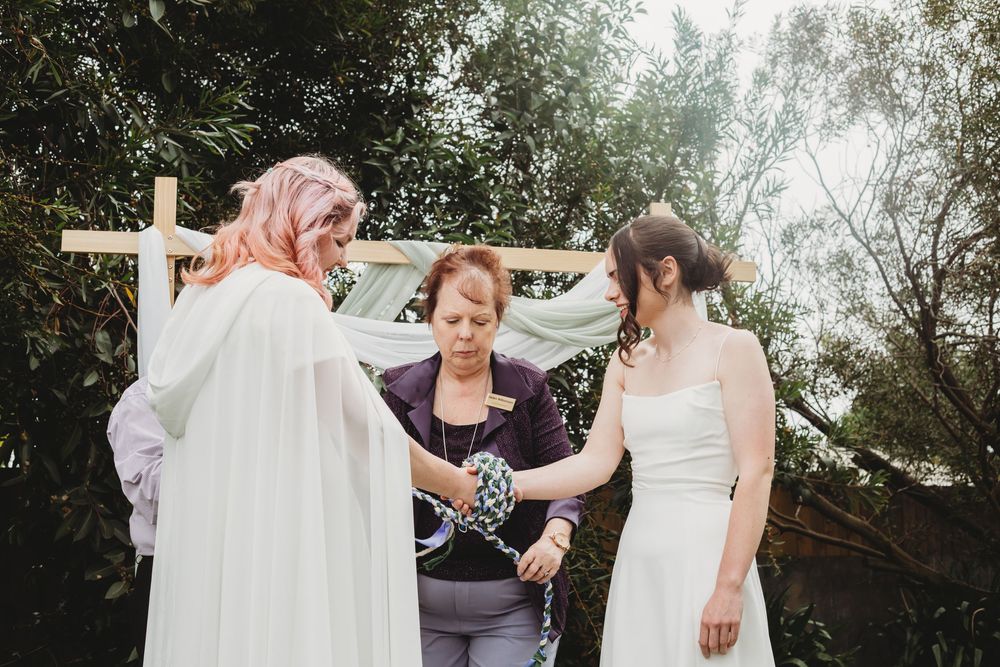 Group in white and purple attire collaborate to set up a wooden wedding arch with flowing fabric outdoors.