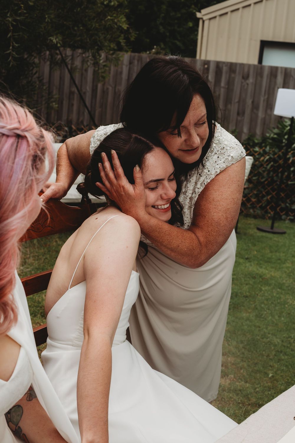 An emotional moment between a bride and woman in gray dress sharing a tender embrace in a backyard garden setting.